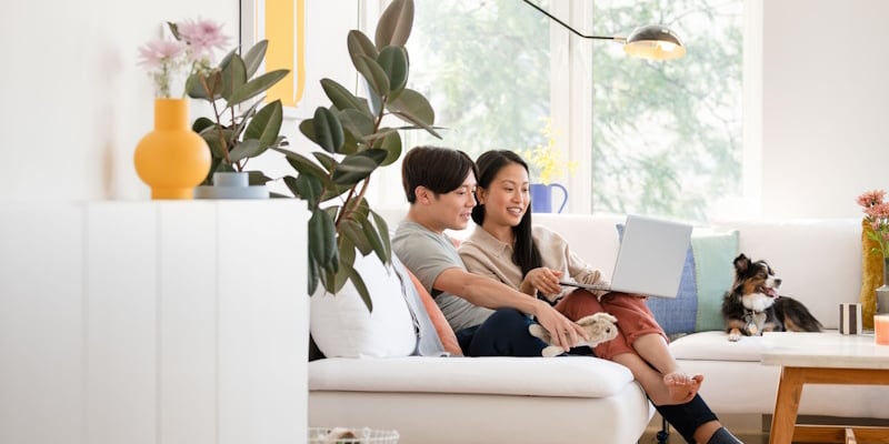 A man and a woman look at a laptop. They are sitting on a sofa in a sunny room